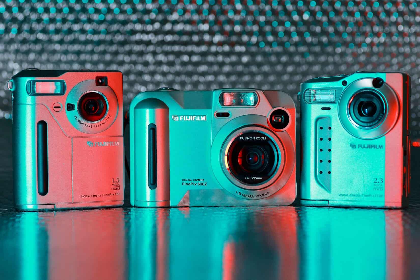 Three vintage Fujifilm digital cameras under colored light